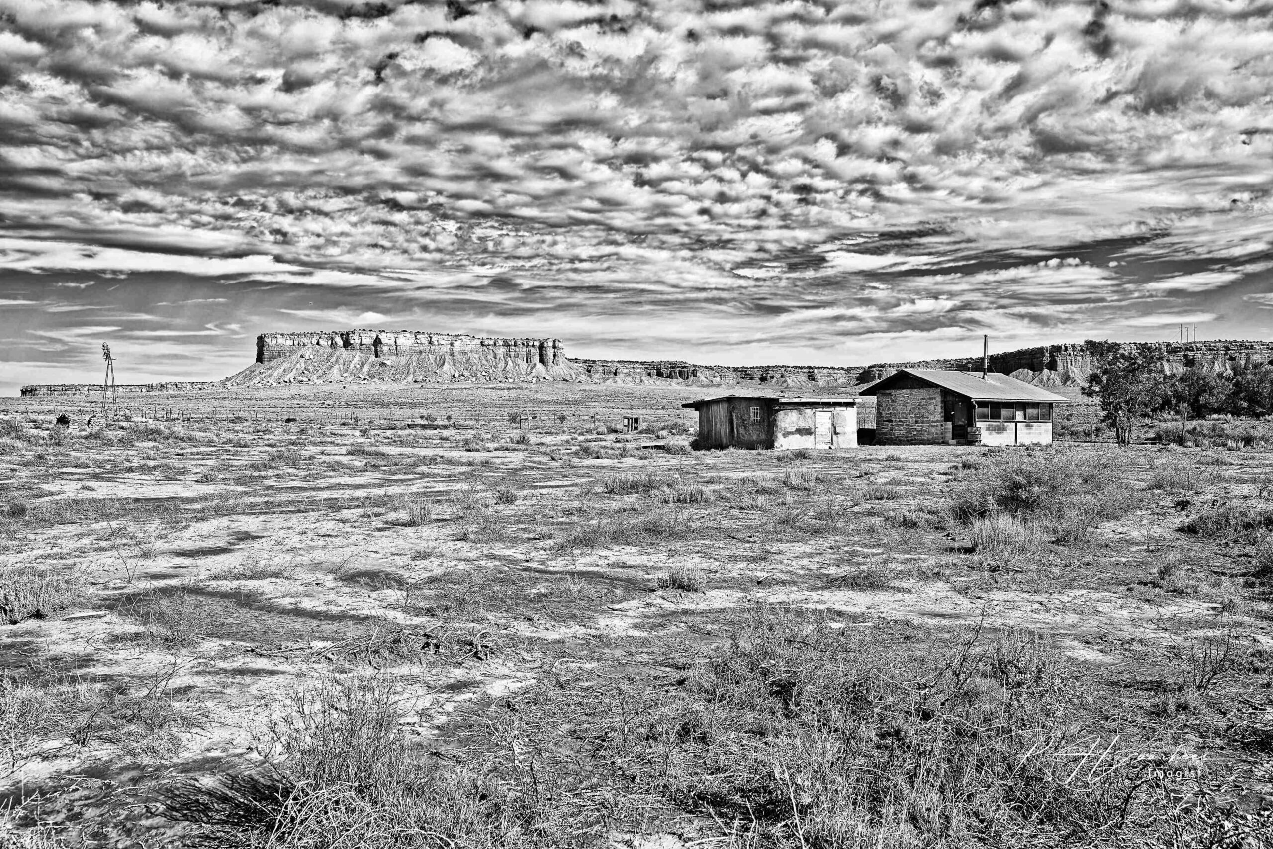 Stone houses and a windmill on open desert below a mesa, under a sky full of clouds